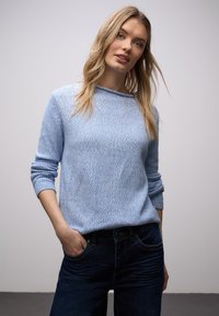 Femme blonde aux cheveux lâchés portant un pull en maille bleu clair et un jean bleu foncé, debout à l'intérieur avec une main dans la poche.