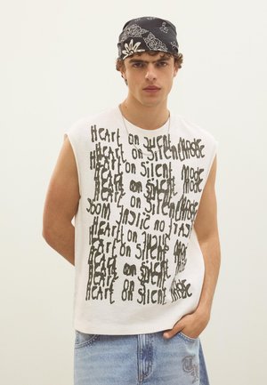 Fiatal férfi fehér ujjatlan ingben, fekete ismétlődő szöveggel és fekete bandanában, rajta fehér virágminták, egy egyszínű háttér előtt áll.