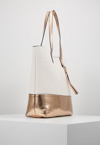 Bolso tote de cuero blanco con una base metálica de color rosa dorado, cierre con cremallera, dos asas y una textura suave. Vista de perfil lateral.