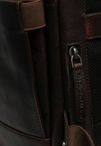 Borsa in pelle marrone con superficie testurizzata, pannello laterale in rete, chiusura con zip e accessori in metallo, e una etichetta in pelle con logo.