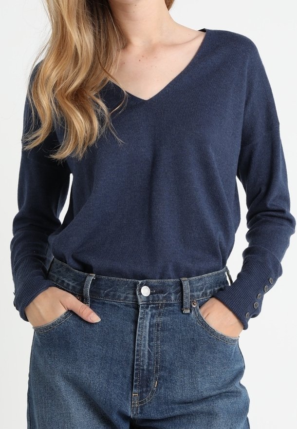 Femme portant un pull bleu marine à col en V et un jean bleu taille haute, les mains dans les poches, sur un fond uni.