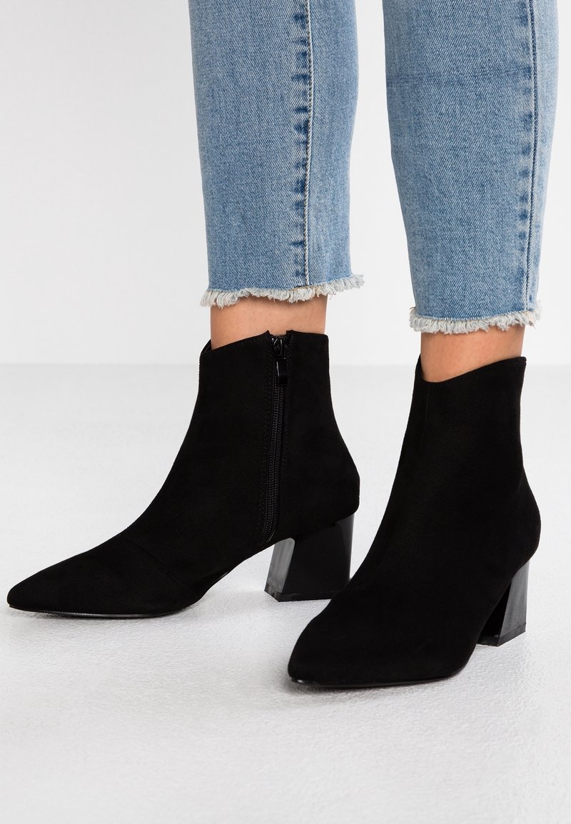 Botas de tobillo de ante negro con punta afilada y tacón cuadrado. Cuentan con un cierre lateral y una textura suave. Se combinan con vaqueros azules con dobladillo deshilachado.