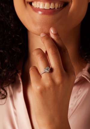 Silver heart-shaped ring with a large central gem, surrounded by smaller stones on the band, worn on a hand with lightly textured skin.