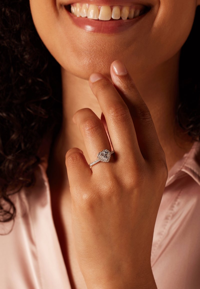 Zilveren hartvormige ring met een grote centrale edelsteen, omringd door kleinere stenen op de band, gedragen aan een hand met lichtgetextureerde huid.