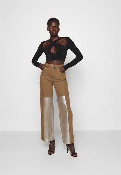 Top corto de manga larga negro con detalle de ojal, combinado con pantalones marrones de cintura alta que presentan paneles plateados brillantes. La modelo está de pie con las manos en las caderas.