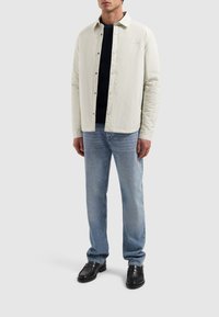 Lichtgekleurde, knoopsluiting jas met een gestructureerde kraag, gedragen over een donkere trui. Combinatie met lichtblauwe jeans en zwarte schoenen.