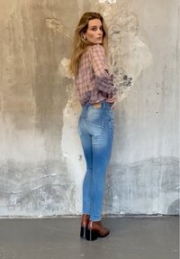 Jeans skinny azzurri chiaro con effetto scolorito, abbinati a una blusa rosa a quadri. Stivali marroni con tacco completano l'outfit. Sfondo neutro.