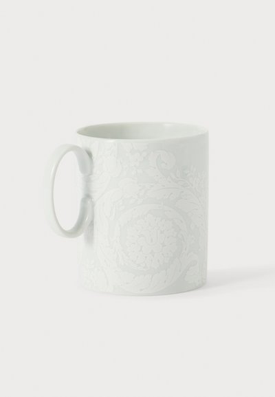 Tasse en céramique blanche avec un motif floral et feuillu délicatement en relief et une anse arrondie sur un fond clair uni.