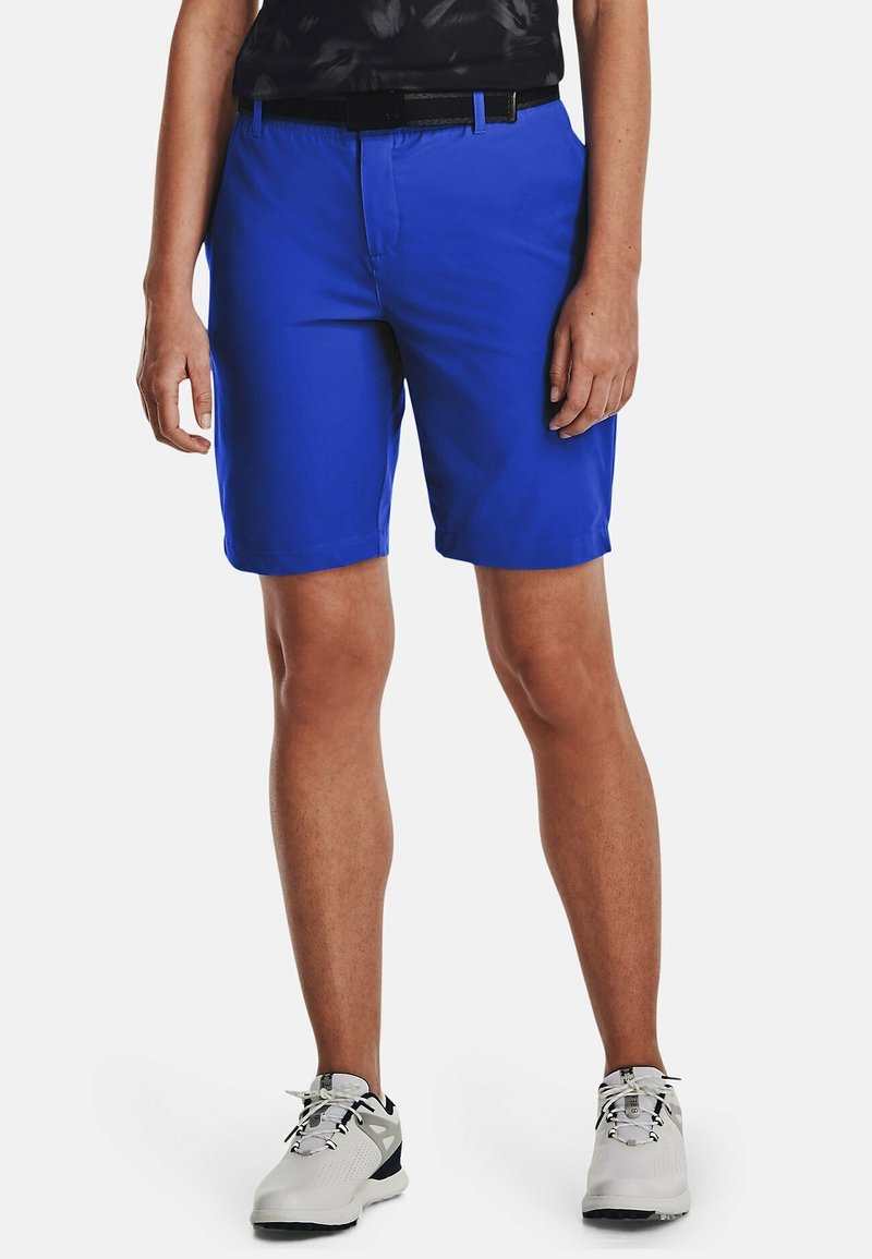 Person som bär ljusblå shorts, svart bälte, svart skjorta och vita träningsskor, stående mot en vit bakgrund.