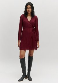 Bordeaux plooien wikkeljurk met lange mouwen en een strikceintuur, met een V-halslijn. Gestyled met kniehoge zwarte laarzen.