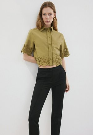 Mujer con una camisa corta verde oliva con botones y detalles de encaje, combinada con pantalones negros de cintura alta, de pie frente a una pared blanca.