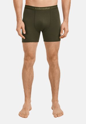 Mand iført olivengrønne boxer briefs med "icebreaker" linning, stående barfodet mod en ensfarvet baggrund.
