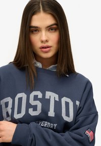 Sudadera de color azul marino con la impresión gris "BOSTON" y "SUPERDRY" debajo. Presenta puños acanalados y un parche con la bandera estadounidense en la manga.