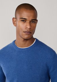 Blå stickad tröja med rund halsringning, tillverkad av mjukt tyg, strukturerad yta, bärs över en vit skjorta. Enkel och ren design.