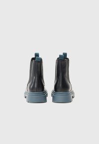 Botas de tornozelo pretas em couro com um acabamento texturizado, solas de borracha azul em contraste e puxadores em tecido na parte de trás para facilitar o uso.