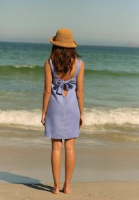 Blaues Leinenkleid mit einem Rückenausschnitt und Schleifendetail, kombiniert mit einem Strohhut. Barfuß auf einem sandigen Strand in der Nähe des Ozeans stehend.