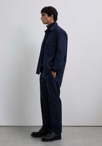 Chaqueta azul marino con dos bolsillos en el pecho, combinada con pantalones a juego. El modelo se encuentra de pie en zapatos negros sobre un suelo texturizado frente a un fondo sencillo.