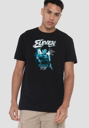 Junger Mann trägt ein schwarzes T-Shirt mit blauem Motiv von Eleven aus Stranger Things, Text „Friends Help Friends“ und „Tales from the Upside Down.“