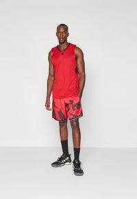 Rotes ärmelloses Sportshirt mit schwarzen Akzenten, kombiniert mit roten Shorts mit einem schwarzen gezackten Muster. Schwarze Sportschuhe mit grünen Details.
