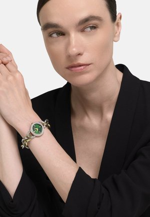 Nő hátratűzött hajjal, fekete blézert visel, kezén zöld számlapú, ezüst és arany színű láncos karóra található.