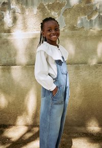 Chemise blanche à manches longues avec col à volants, associée à une salopette en jean bleu clair avec poches et bretelles réglables.