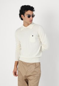 Sweater de malha creme com decote tipo colarinho, apresentando um bolso no peito com um botão preto. Combinado com calças castanhas claras.