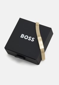 Aukso pynimo apyrankė su stačiakampiu užsegimu, pateikta juodame dėkle, kuriame baltomis raidėmis užrašyta "BOSS". Sklandi tekstūra, modernus dizainas.