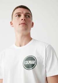 Camiseta blanca de algodón con un logo redondo de camuflaje verde y el texto "COLMAR" en blanco. Diseño clásico de cuello redondo, mangas cortas.