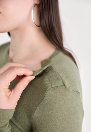 Long-sleeve, olive green sweater with a soft texture. Features a scoop neck and a stitched shoulder detail, with a hand grasping the collar.