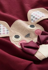 Bordeauxfarbener Pullover mit einem beigen Tiergesicht-Applikation, karierter Ohren, gestickten Details und einem dunkelvioletten Schleifenakzent.