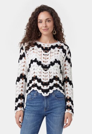 Donna con capelli ricci che indossa orecchini d'oro, top a maniche lunghe lavorato all'uncinetto bianco e nero e jeans blu, in piedi su uno sfondo bianco.