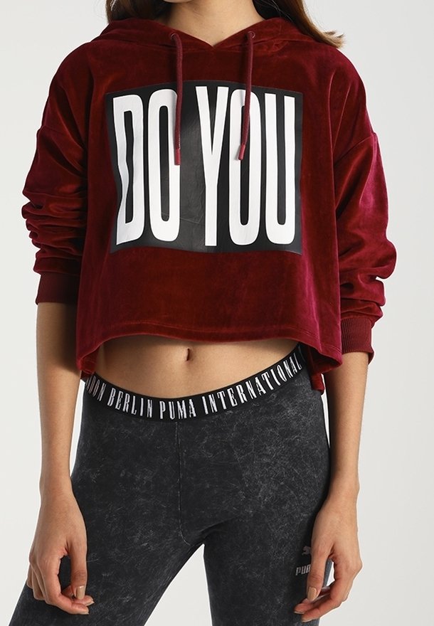 Femme portant un sweat à capuche court rouge avec le texte "DO YOU" et des leggings noirs Puma avec une ceinture marquée, debout les mains détendues.