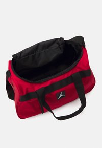 Bolsa de deporte roja hecha de un tejido duradero, con una cremallera negra, correas negras y un parche con un diseño de silueta. El interior es negro.