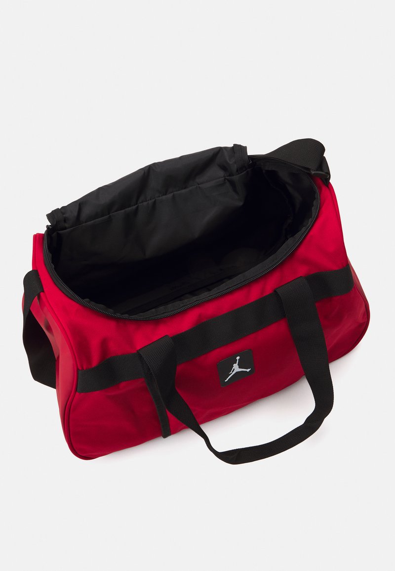 Borsa duffel rossa realizzata in tessuto resistente, con una cerniera nera, manici neri e una toppa con logo dal design silhouette. L'interno è di colore nero.