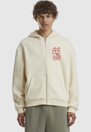 Krém színű, cipzáros kapucnis pulóver, puha anyagból készült, bal mellen piros macska grafikával. Elöl zsebekkel és laza szabással rendelkezik.