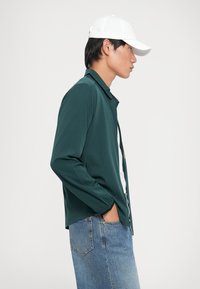 Σκούρο πράσινο πουκάμισο με μακριά μανίκια και γιακά, σε συνδυασμό με ανοιχτού μπλε τζιν. Λευκό καπέλο φορεμένο αντίστροφα. Λείο υφή, χαλαρός σχεδιασμός.