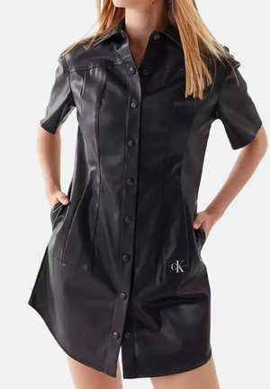Frau trägt ein kurzärmeliges schwarzes Lederknopfkleid mit Taschen und kleinem Calvin Klein-Logo vorne unten.