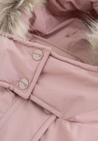 Veste isolante rose avec une capuche doublée de fourrure douce, dotée de boutons ronds portant l'inscription "UNLIMITED TEC" et d'un tissu lisse et texturé.
