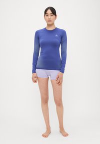 Αθλητικό μπλουζάκι με μακριά μανίκια σε έντονο μπλε χρώμα, με διακριτικό σχεδιασμό διαβάθμισης. Συνδυάζεται με ανοιχτό μωβ σορτς, που αναδεικνύει εφαρμοστή γραμμή.