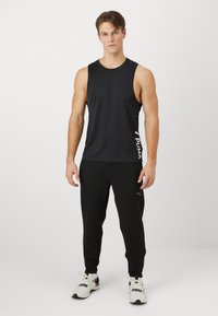 Zwarte mouwloze sportshirt met PUMA-logo en bijpassende zwarte broek met structuur. Model draagt witte sportschoenen met zwarte accenten.