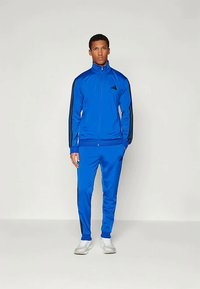 Mėlynas sportinis kombinezonas, pagamintas iš lygaus audinio, su juodomis šoninėmis juostelėmis ant striukės ir kelnių, su užtrauktuku prie kaklo ir logotipu ant krūtinės.