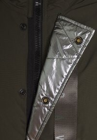 Olijfgroen jack met een zwarte rits, zilverkleurig doorgestikt paneel en gouden knopen, met textuurdetails in de stof.