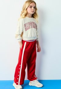 Creme sweatshirt med teksten "HAPPINESS", parret med røde sweatpants med hvide side striber og knaplukninger. Hvide sneakers fuldender looket.