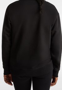 Person som bär en enkel svart långärmad tröja och svarta byxor, sedd bakifrån med flätat hår som hänger ner.