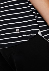 Hém de chemise à rayures noires et blanches avec une petite étiquette métallique ronde, associé à un pantalon noir et un bras visible reposant sur le côté.