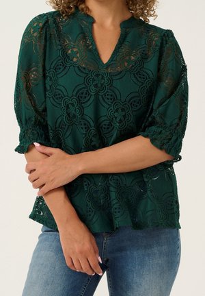 Vrouw draagt een donkergroene kanten blouse met bloemmotieven en driekwart mouwen, gecombineerd met een blauwe spijkerbroek.