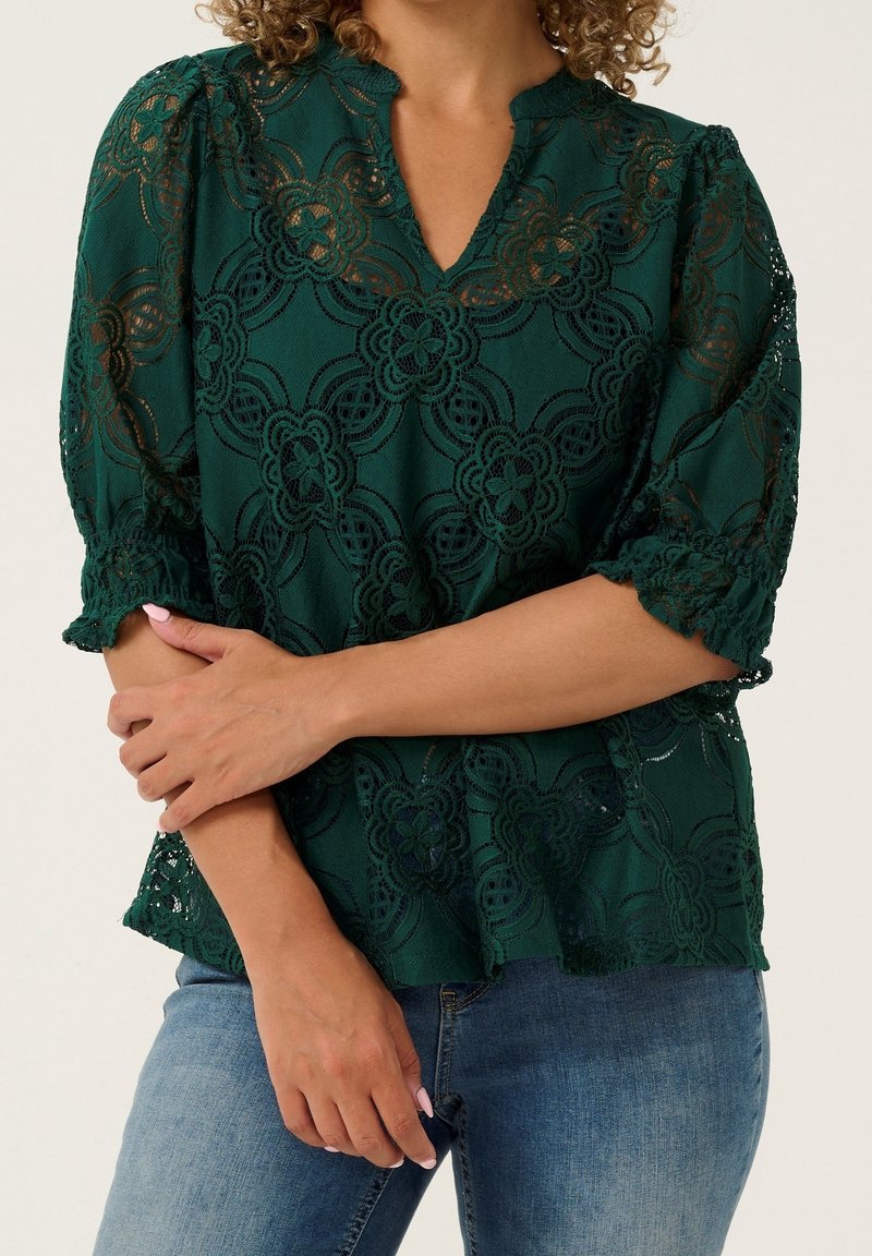 Femme portant un chemisier en dentelle vert foncé avec des motifs floraux et des manches trois-quarts, associé à un jean bleu.