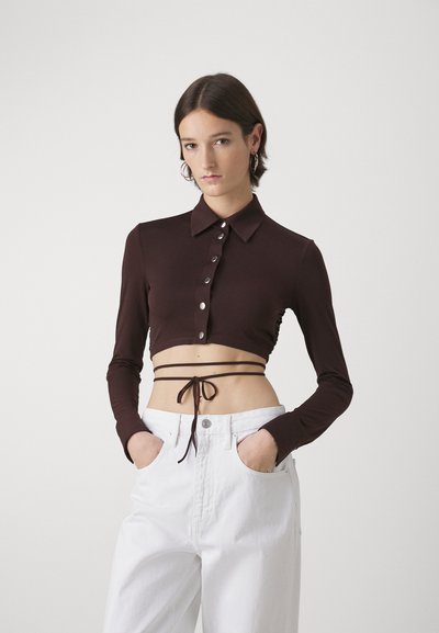 Brown, cropped long-sleeve shirt with a collar and button closure, featuring side ruching and a tie detail around the waist. Paired with white trousers.