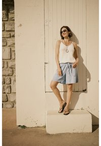 Lightweight, knitted white tank top, blue drawstring shorts, black flip-flops, and sunglasses, posed against a neutral wall with wooden door.