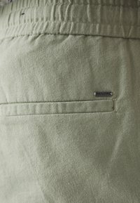 Grüner Stoff mit einer weichen Textur, ausgestattet mit einer Rückentasche und einem kleinen Markenetikett an der Seite, mit sichtbaren Nähdetails.